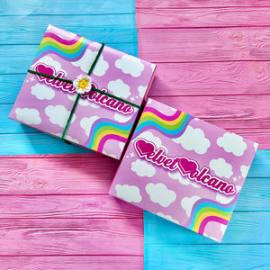 Two pastel pink boxes with white cloud pattern and wiggly pastel rainbow accents in the upper left and bottom right. One box has a crochet flower tie wrapped around it. The boxes are on a pink and turquoise tinted wood background